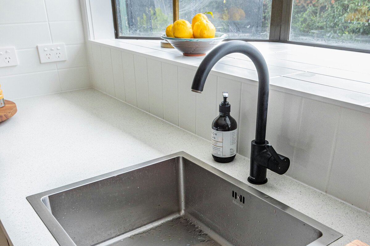 Black mixer tap and stainless steel sink on a stone benchtop in an Australian kitchen, where an under-sink water filter sits in the cupboard below